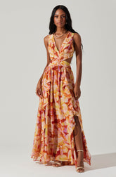 A woman is wearing a sleeveless, V-neck maxi dress with a floral pattern in shades of pink, orange, yellow, and red. The dress features cut-out sides at the waist and tiers of ruffles down the skirt, which has a high slit on one side. She is accessorized with a gold necklace, rings, and open-toe heels.